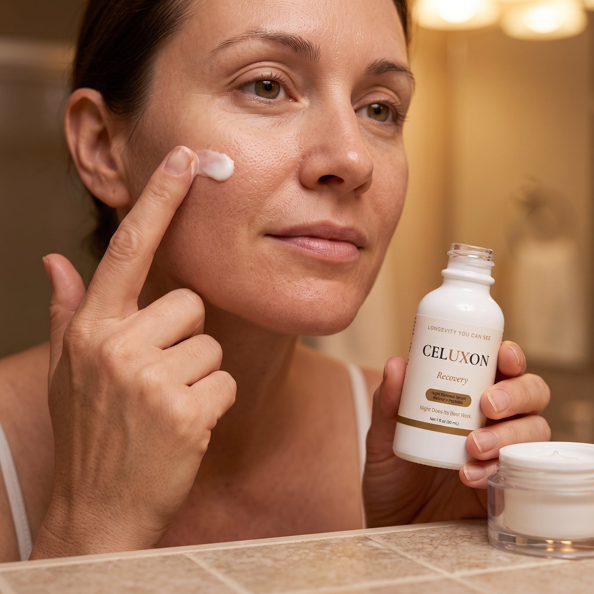 A woman in a bathroom applies a white cream to her cheek while holding a bottle of CELUXON Recovery facial cleanser and a small jar on the counter.