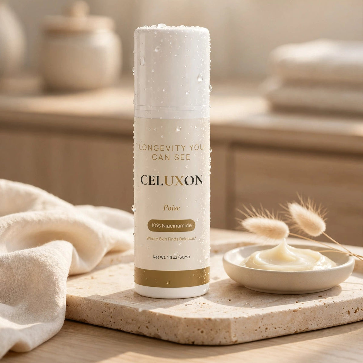 A white cylindrical skincare bottle labeled CELUXON with gold text, standing on a stone tray beside a small dish of cream and dried grass stems in a softly lit bathroom setting.